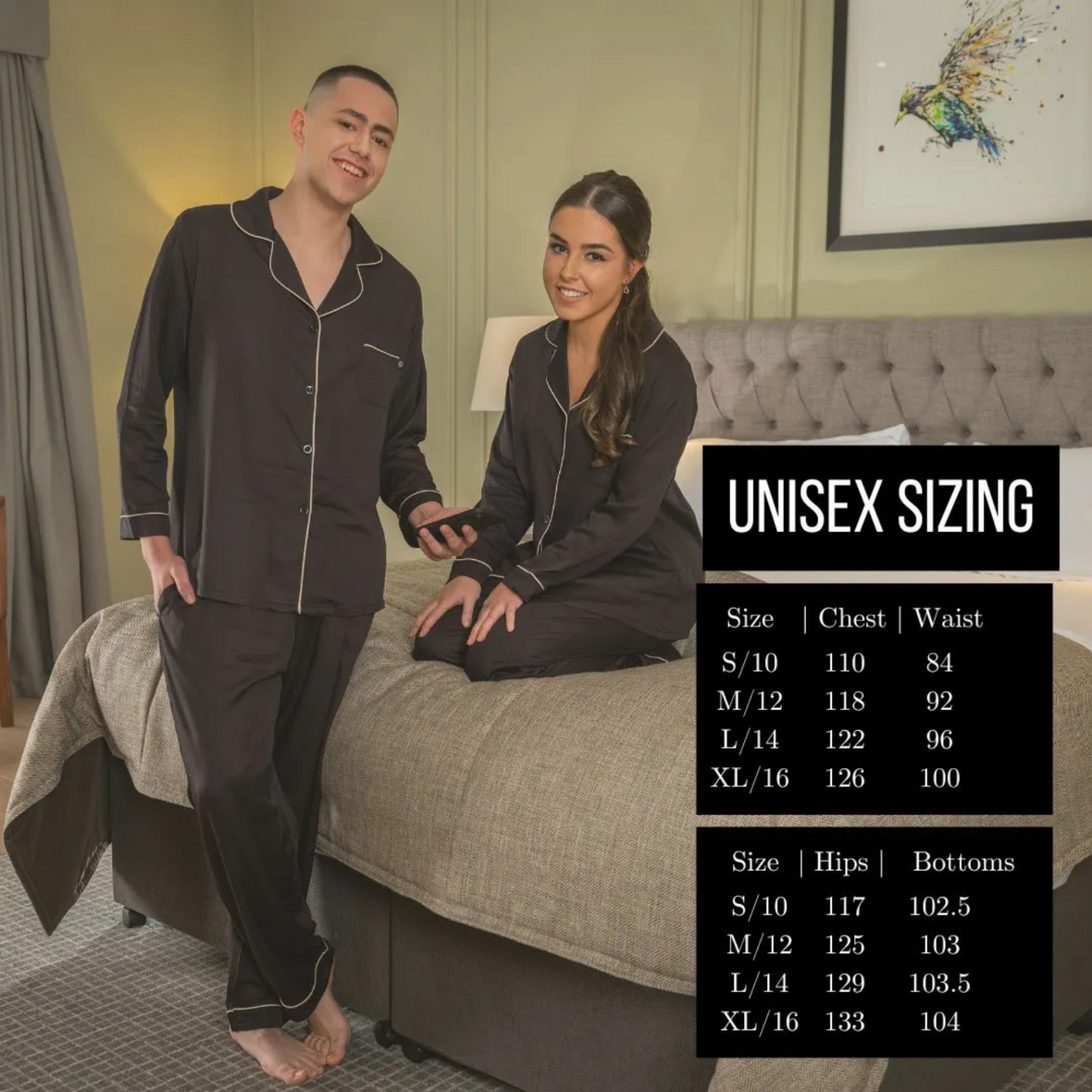 A man standing while a woman is sitting, both wearing  the Jo Browne Luxury Unisex Bamboo Pyjamas. A unisex size guide for the pyjamas is included in the image. 