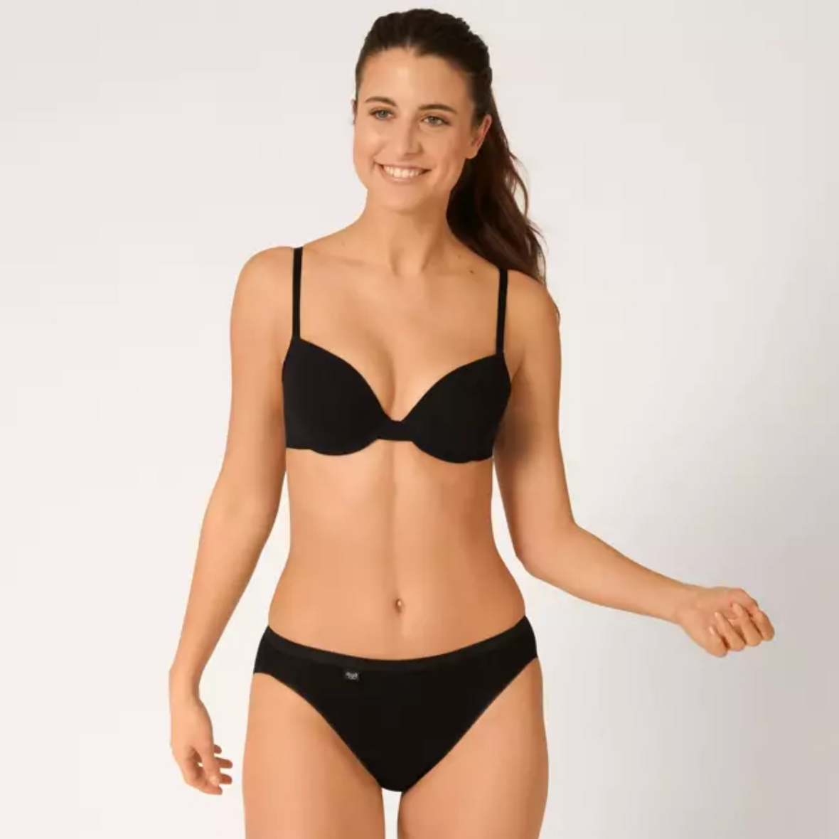 A model wearing the Sloggi Tai Brief in Black looking to the left and smiling with her right arm raised.